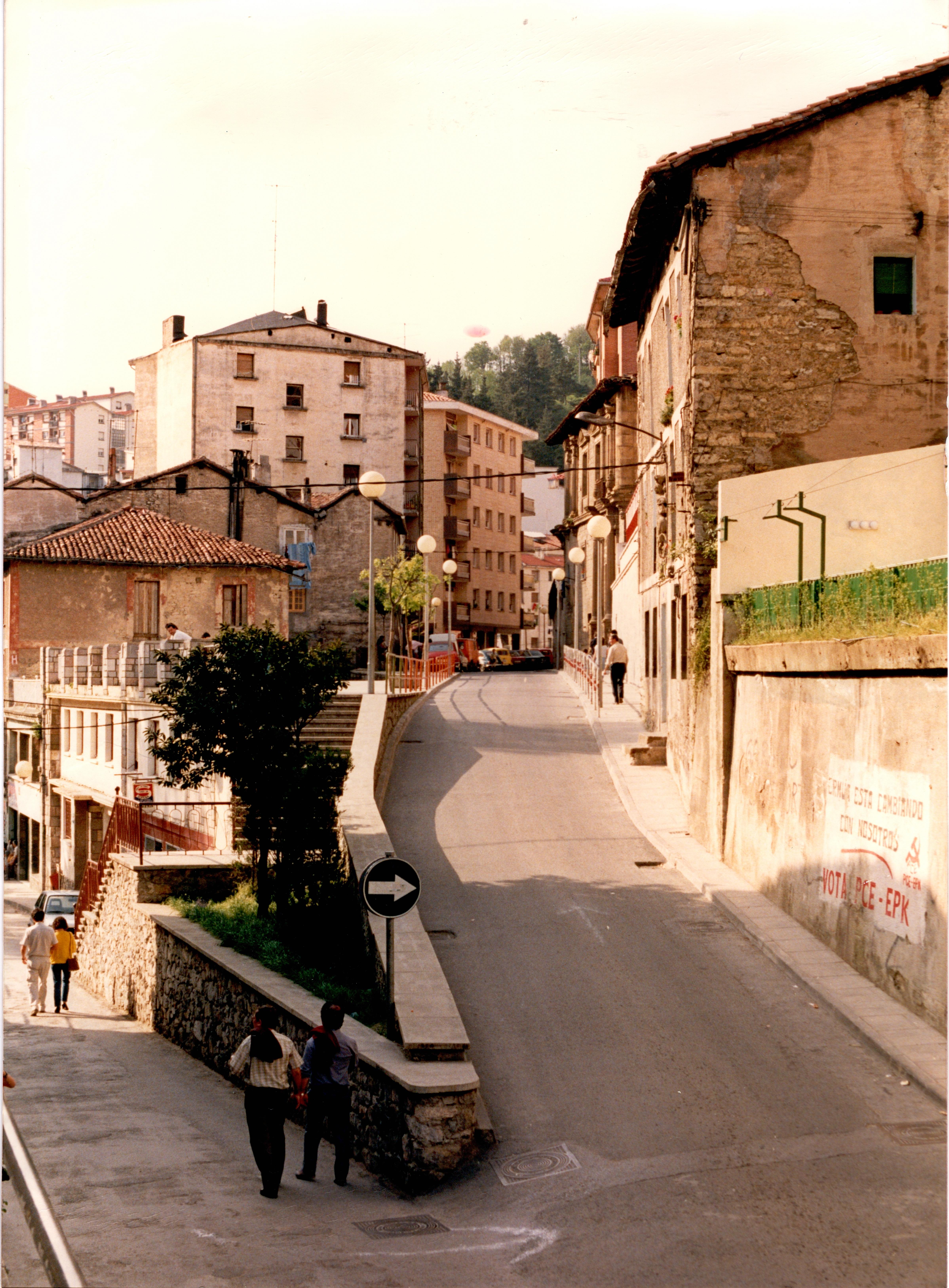 Se muestra la calle Goienkale y a la izquierda la calle Izelaieta. Se separan dos calles, la calle de la izquierda parece peatonal y se ven algunos edificios. Por esa calle bajan dos parejas. A la derecha, en la calle Goienkale se muestra una pequeña cuesta y la calle está despejada, en la acera, en el fondo de la fotografía hay algunos peatones.