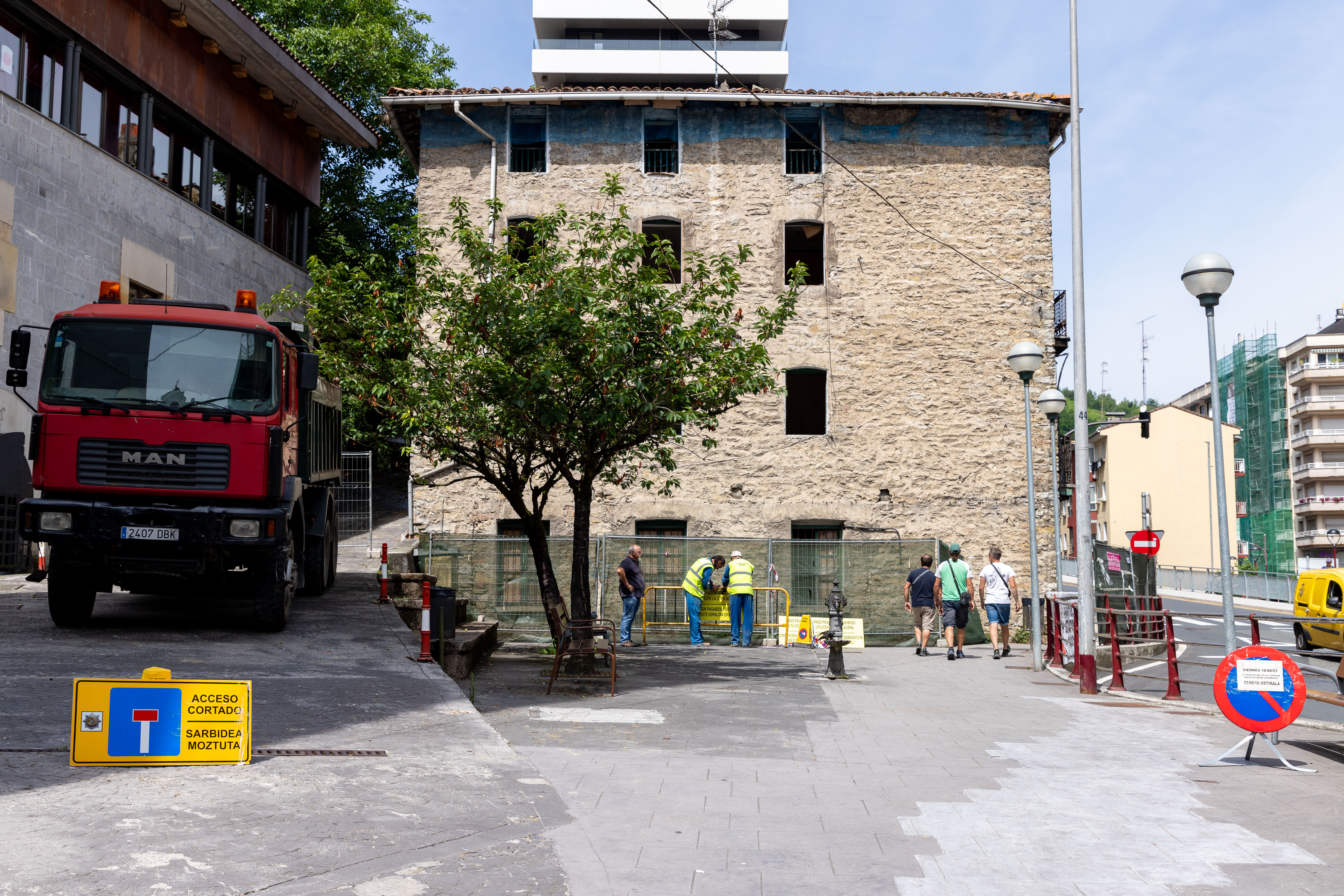 Se ve en el centro de la fotografía el edificio que derribarán. A la izquierda se ve parte de la fachada principal de otro edificio (Torreta) de fachada gris de piedra y algunas ventanas en la misma altura de forma horizontal. Debajo, a pie de calle hay un camión rojo que trabaja en las labores del derribo. Se ven señales que indican que el acceso está cortado. En otra señal se indica que a partir de las 8:00 horas del viernes 16/07/23 no se podrá ir a pie por la zona del derribo. Hay tres trabajadores.