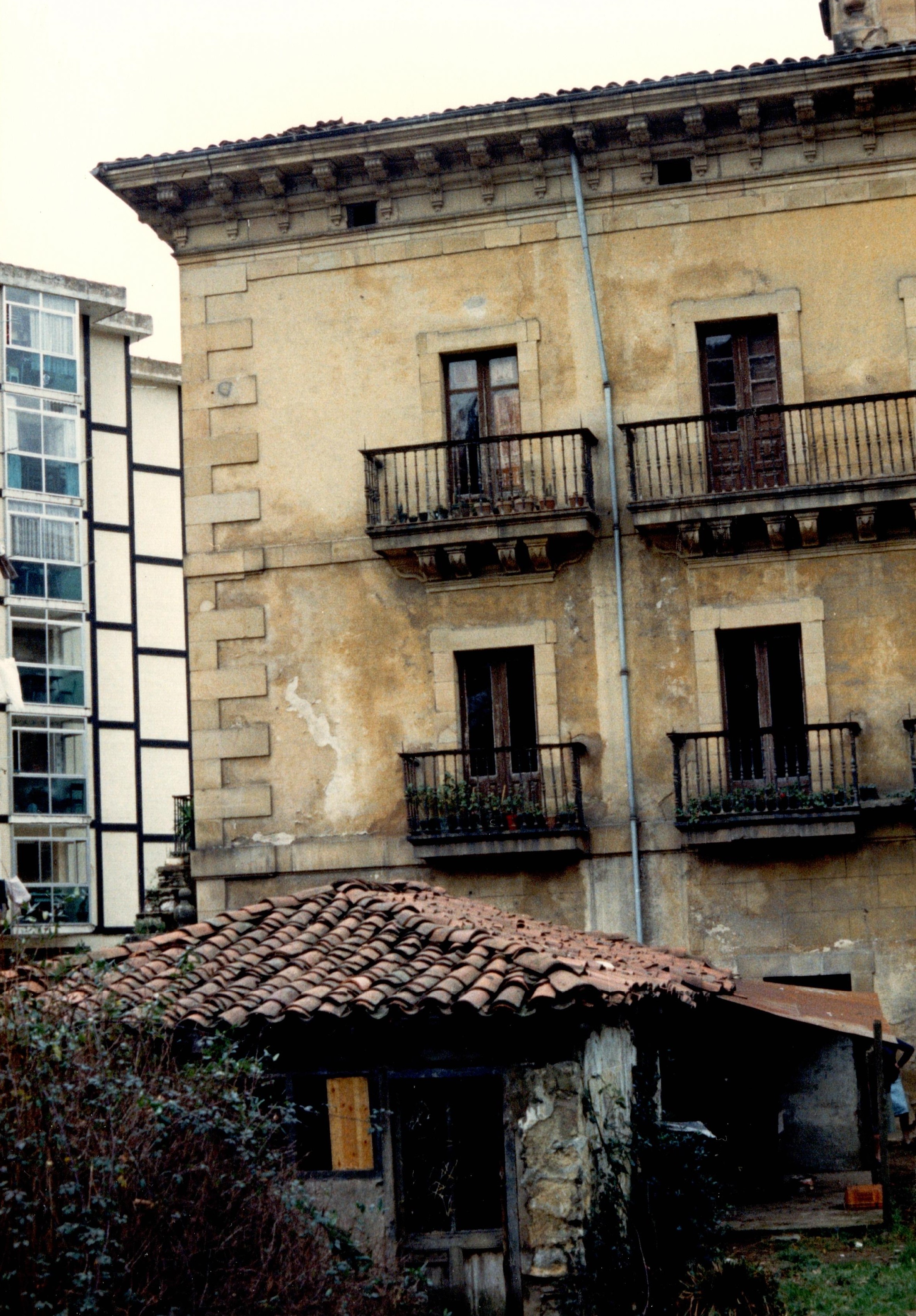 Se ve un edificio de color arena clarita, con cuatro balcones. A la izquierda hay un edificio moderno, un bloque de viviendas con ventanas y fachada blanca con líneas negras. En el interior del jardín, se ve una parcela con hierba verde y una pequeña construcción con techo de ladrillo que podría servir para guardar herramientas o animales.