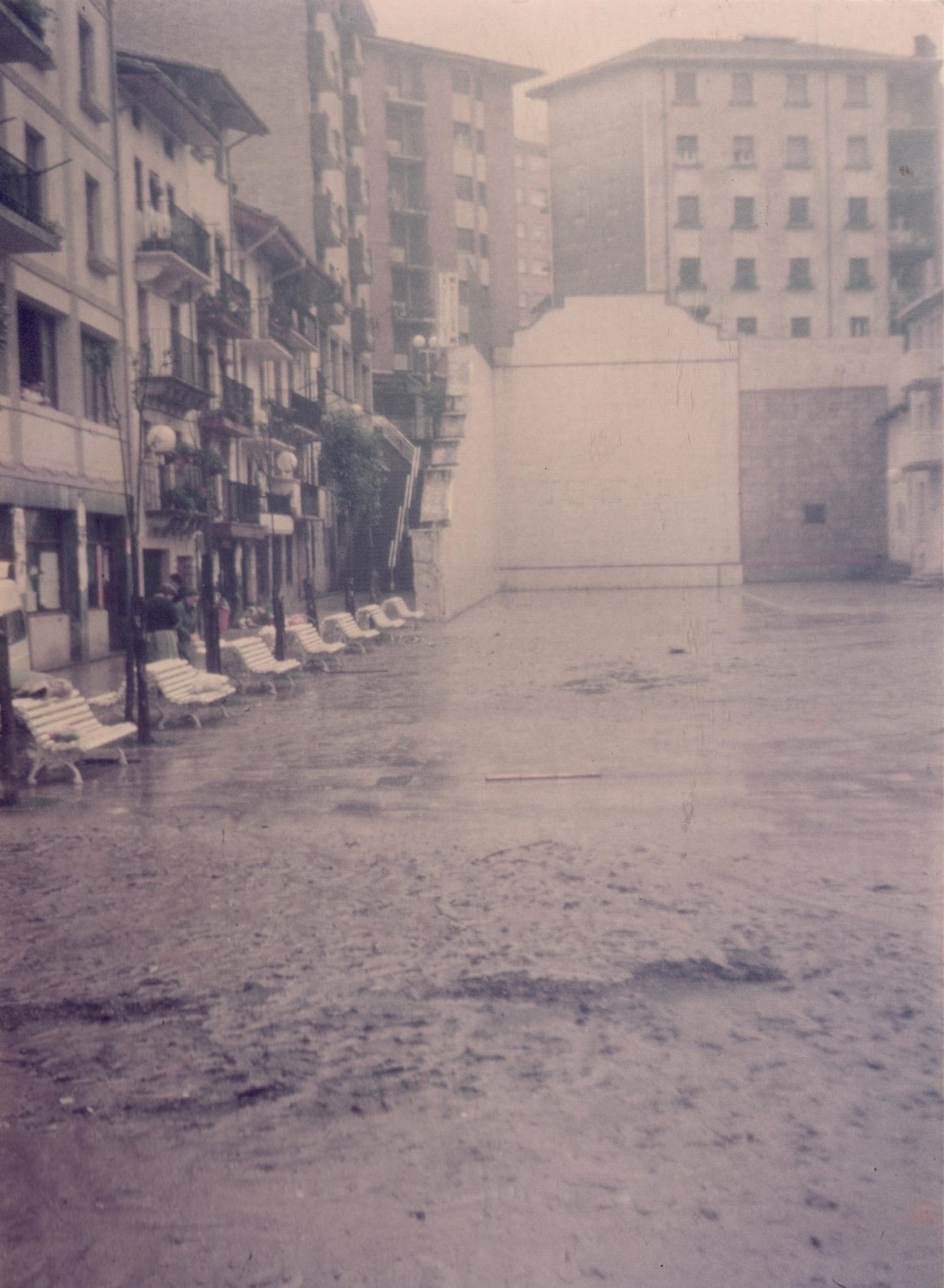 Se ve la plaza Cardenal Orbe y de fondo el frontón. A la izquierda hay siete bancos blancos. Parte del asfalto está perjudicado por los efectos de las inundaciones. A la izquierda hay viviendas y al fondo detrás del frontón también. A la izquierda del frontón se ven unas escaleras que suben y que tienen una barandilla blanca.