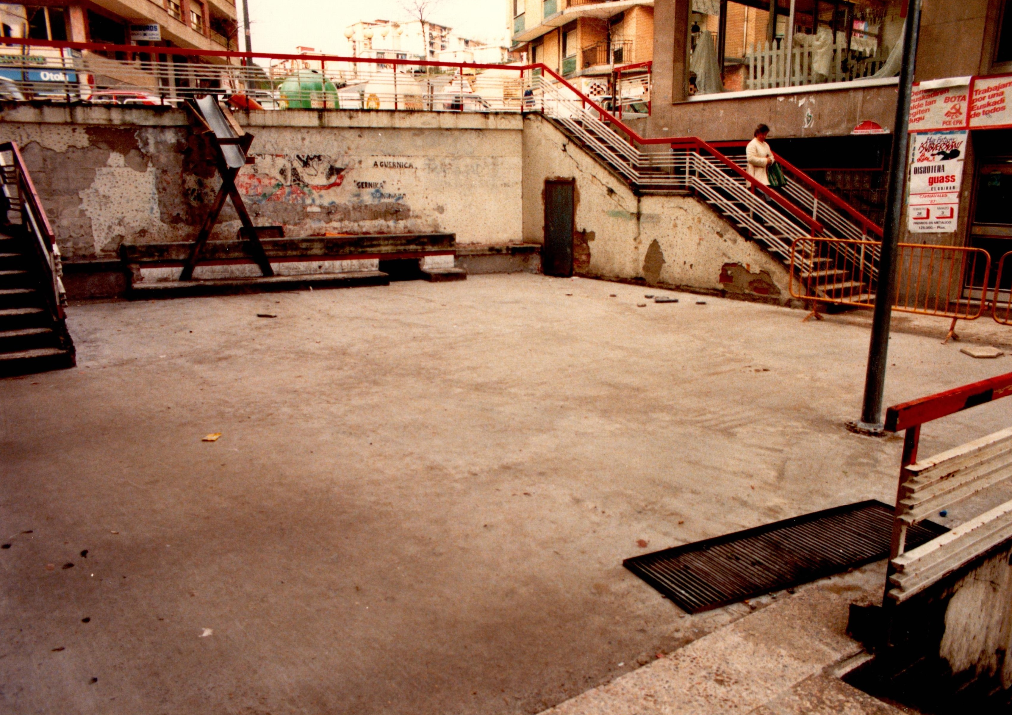 Se muestra una plaza prácticamente vacía con escaleras a los lados. Hay un banco en la parte del fondo y una estructura metálica.